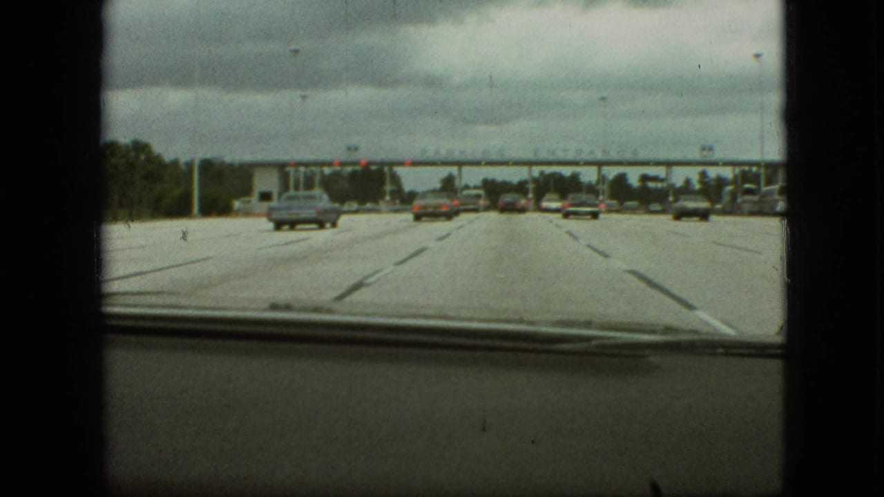 From inside a moving car, late-1960s traffic approaches a broad parking entrance structure in Branson, Missouri, with cloudy sky and dark 8mm frame borders visible.