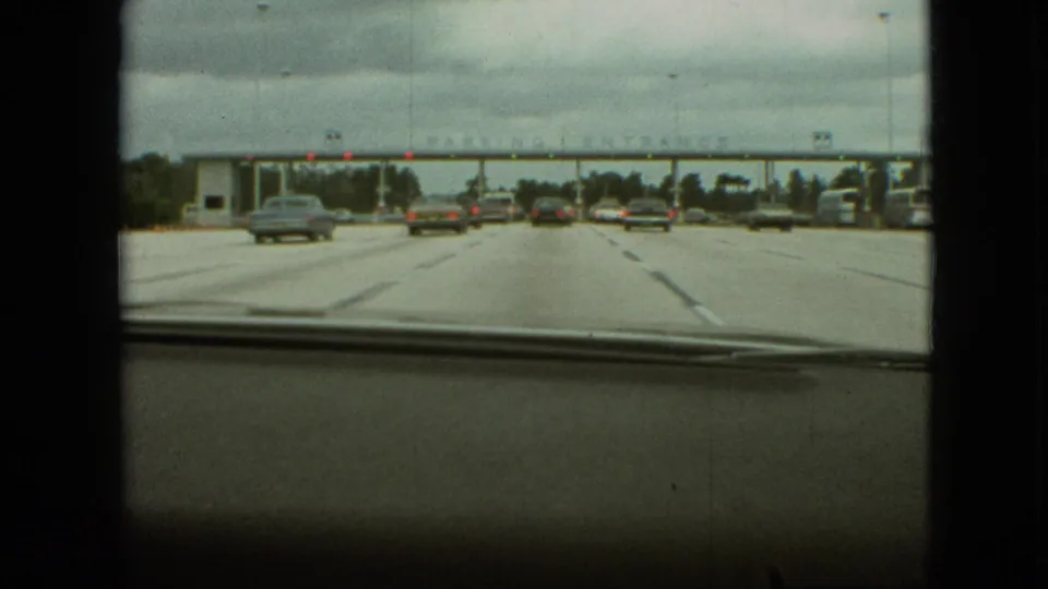 The windshield view shows several automobiles spread across wide lanes as the car nears an overhead parking entrance in the supplied 1968 Branson setting.