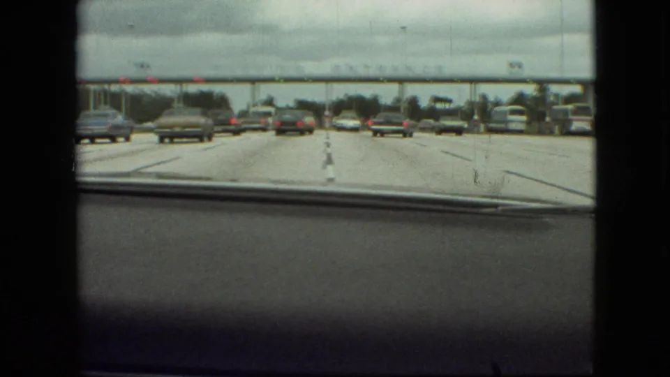 A closer windshield view shows multiple cars in separate lanes approaching the overhead parking entrance structure, with roadside trees and the car dashboard edge visible.