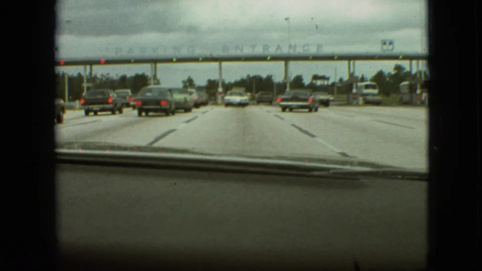 The car approaches beneath an overhead structure where faint parking entrance lettering is visible, with traffic ahead, red lights, and 8mm film borders framing the scene.