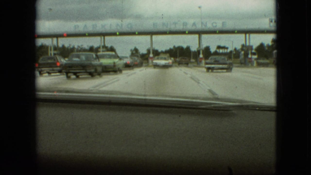 From inside a moving car in Branson, Missouri, 1968, period automobiles approach a broad parking entrance canopy with lane booths visible ahead.