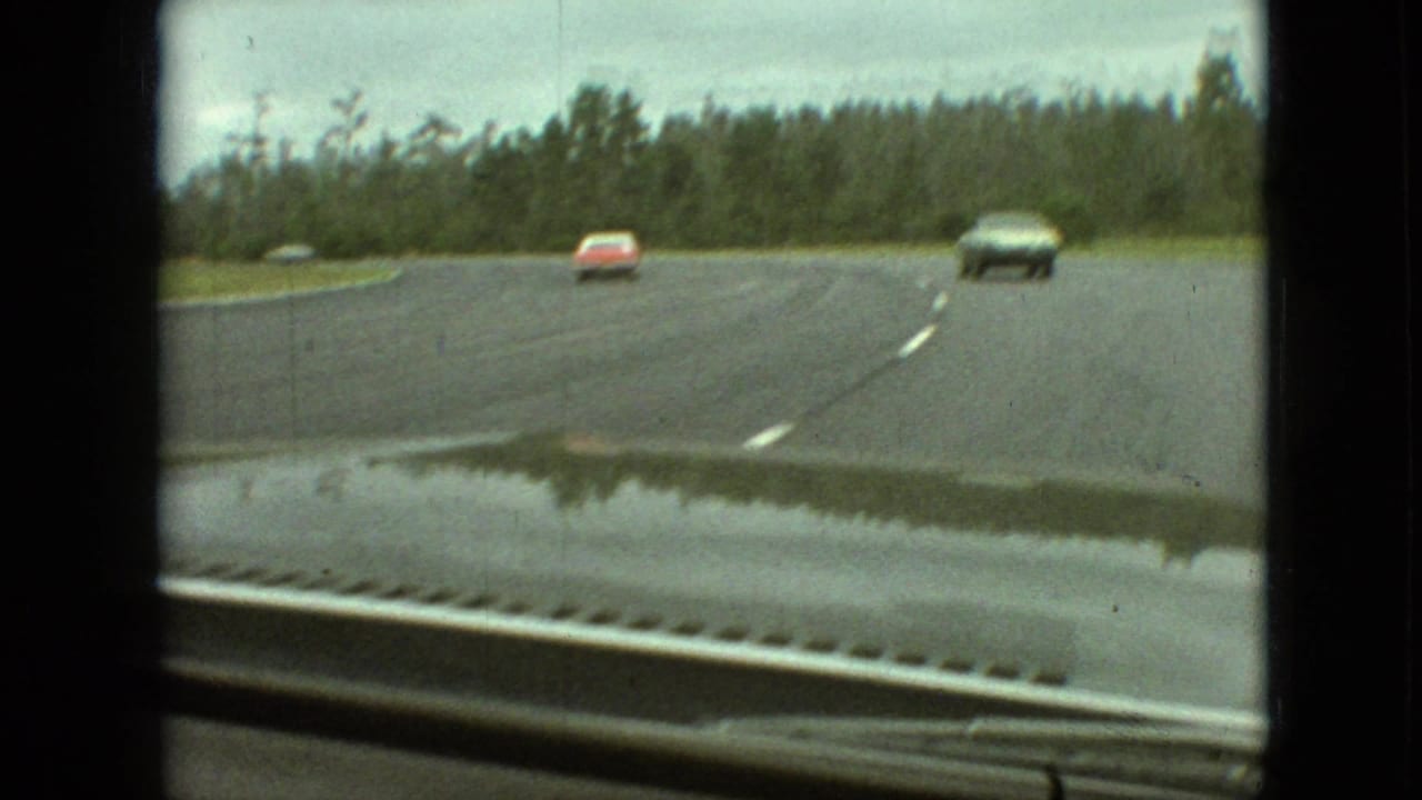 1968 Branson, Missouri windshield view of a rain-darkened curving road bordered by trees, with a red car and a dark sedan approaching from the opposite direction.