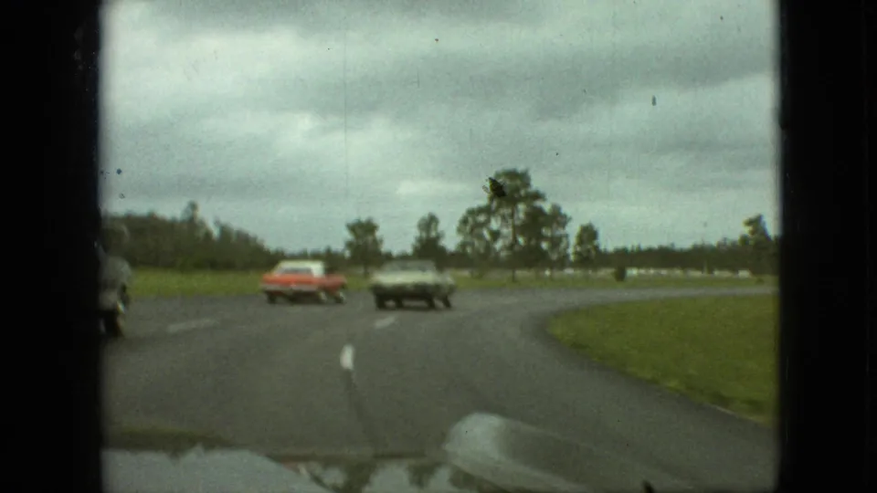 1968 Branson, Missouri road scene from a moving vehicle, with a red car and dark sedan passing along a wet curve bordered by grass and trees.
