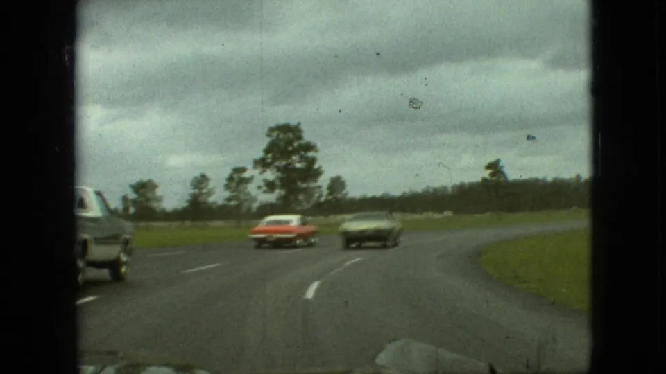 1968 Branson, Missouri home-movie frame showing traffic on a wet curving road, including a red car, a dark sedan, and a pale vehicle at the left edge.