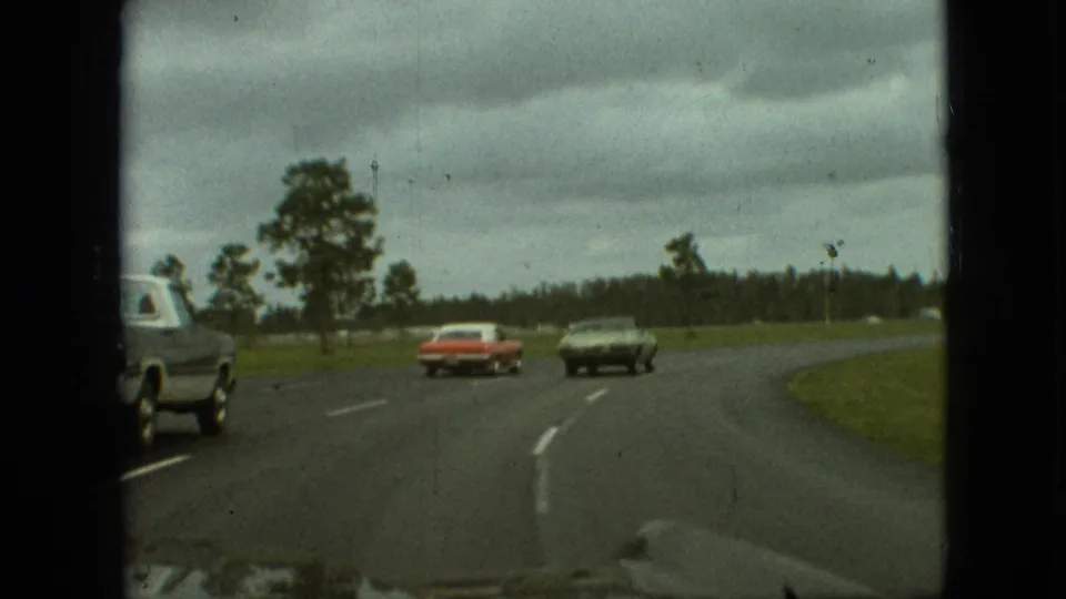 1968 Branson, Missouri home-movie frame showing a red sedan and dark coupe ahead on a curving two-lane highway, with a partial vehicle at the left edge and trees beyond the road.
