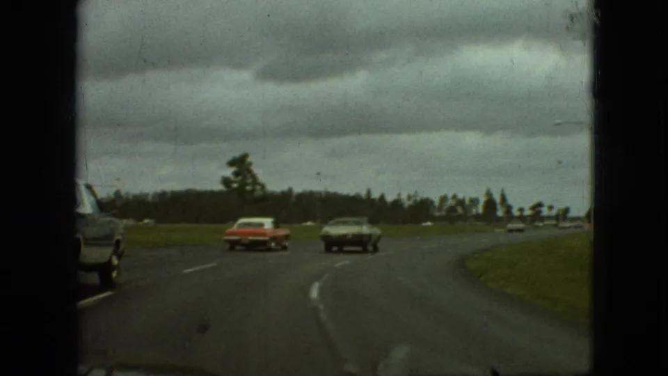1968 Branson, Missouri frame from a moving vehicle, with a red sedan and dark coupe traveling ahead on a curving paved road bordered by grass and distant trees.