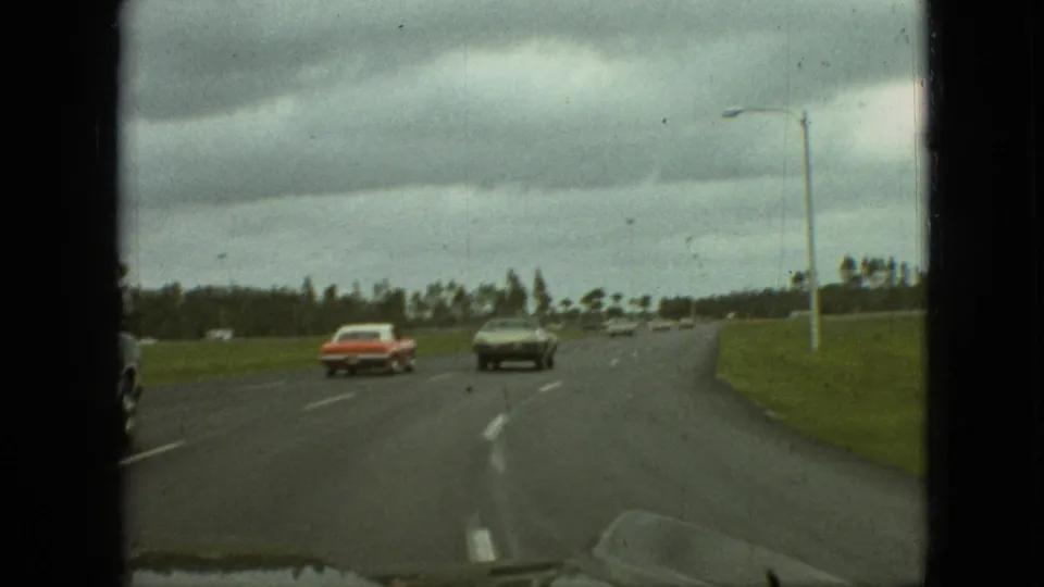 1968 Branson, Missouri highway view showing the same cars farther ahead on a broad curving roadway, with a roadside pole, grassy verge, distant trees, and visible 8mm frame edges.