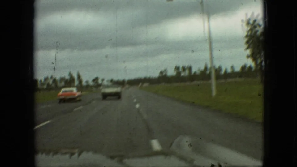 Final 1968 Branson, Missouri frame with the red sedan and dark coupe receding on the road as the passenger-shot view catches the roadside grass, tree line, pole, and dark film borders.