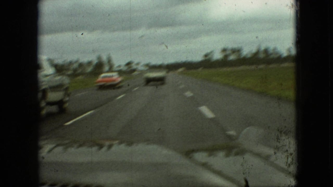 1968 Branson, Missouri 8mm windshield view of a highway with a red car and another vehicle ahead beneath heavy cloud cover.