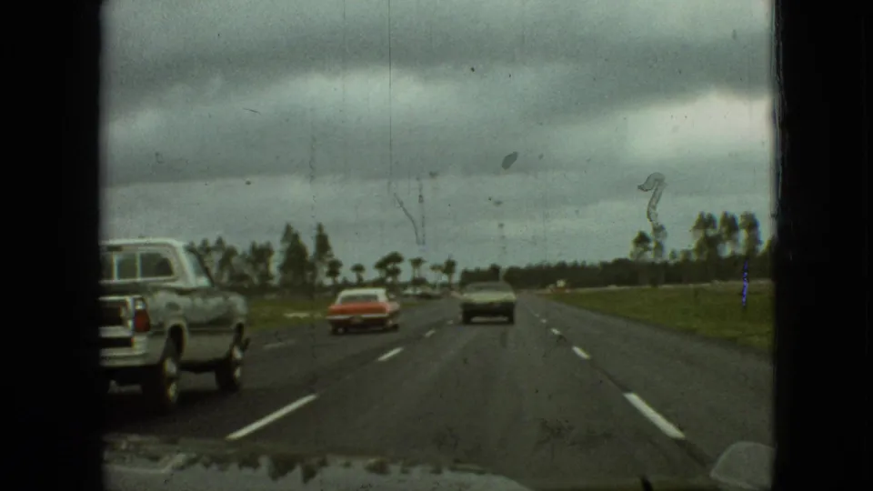 1968 Branson, Missouri 8mm travel frame with a light-colored pickup truck beside the camera car, a red car ahead, and dark clouds over the road.