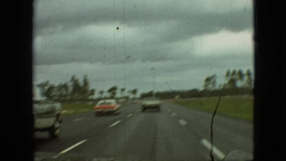 1968 Branson, Missouri 8mm frame showing a soft-focus windshield view of cars traveling on an open highway bordered by grass and distant trees.
