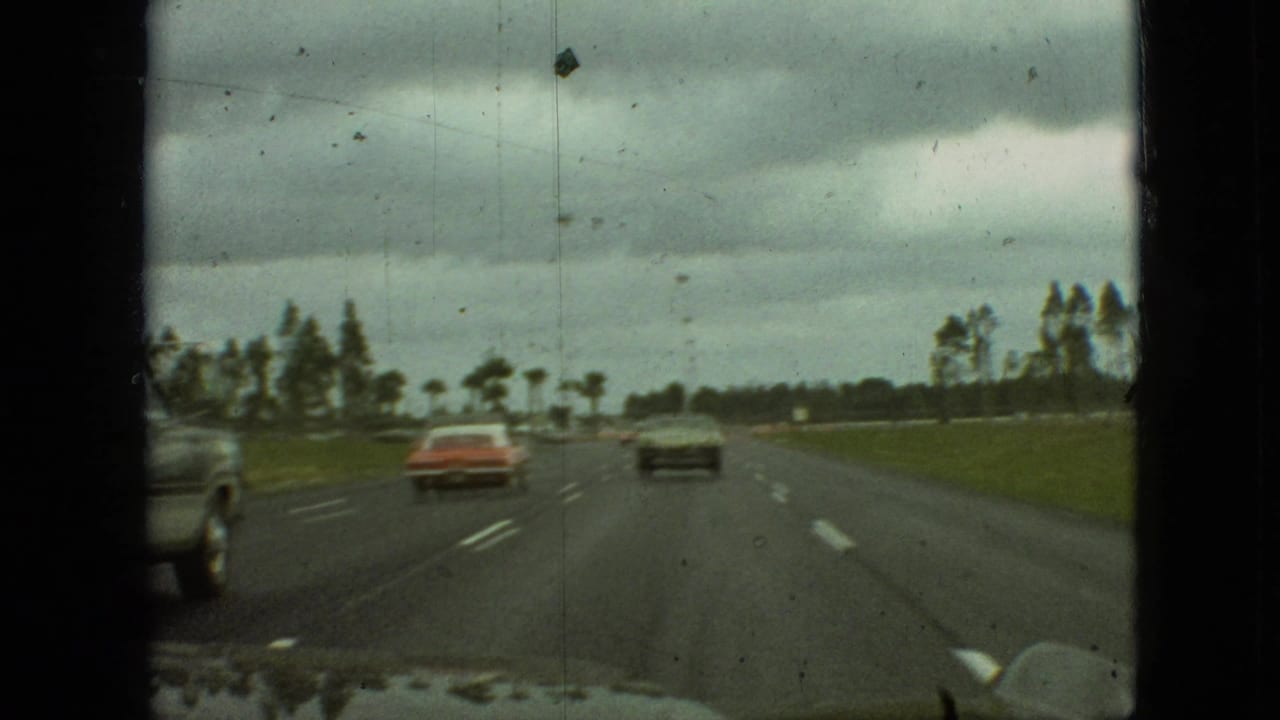 1968 home-movie frame from Branson, Missouri, showing a passenger-side view down a multi-lane highway with a red sedan, a dark sedan, grass, and trees under overcast clouds.