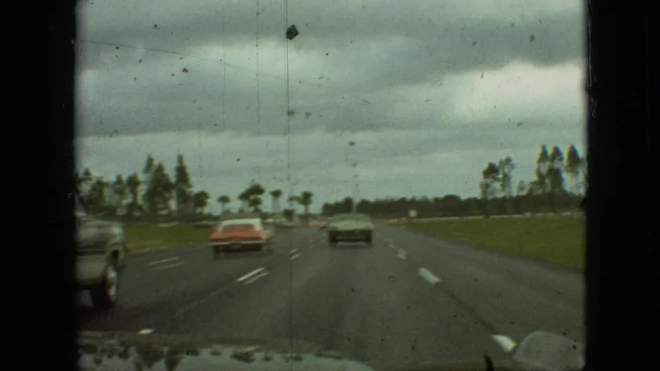 1968 home-movie frame from Branson, Missouri, showing a passenger-side view down a multi-lane highway with a red sedan, a dark sedan, grass, and trees under overcast clouds.