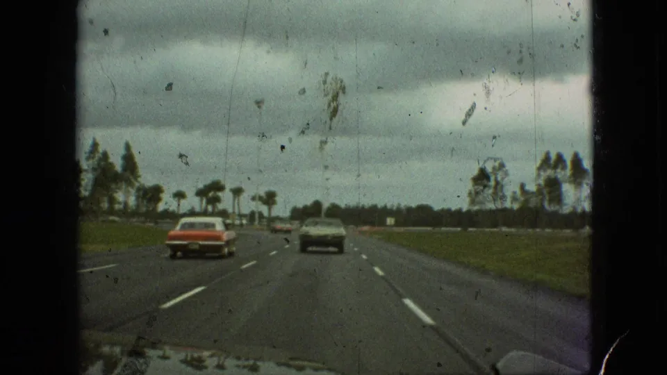 1968 home-movie highway view in Branson, Missouri, with a red sedan and darker car continuing ahead on wet-looking pavement beneath a gray sky.