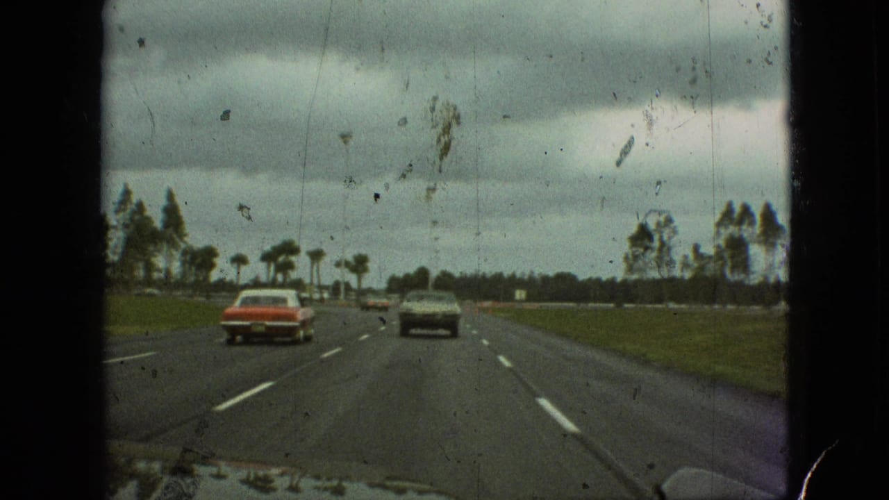 1968 Branson, Missouri 8mm home-movie frame showing a red sedan and a darker car on a multi-lane road with grassy shoulders and roadside trees.