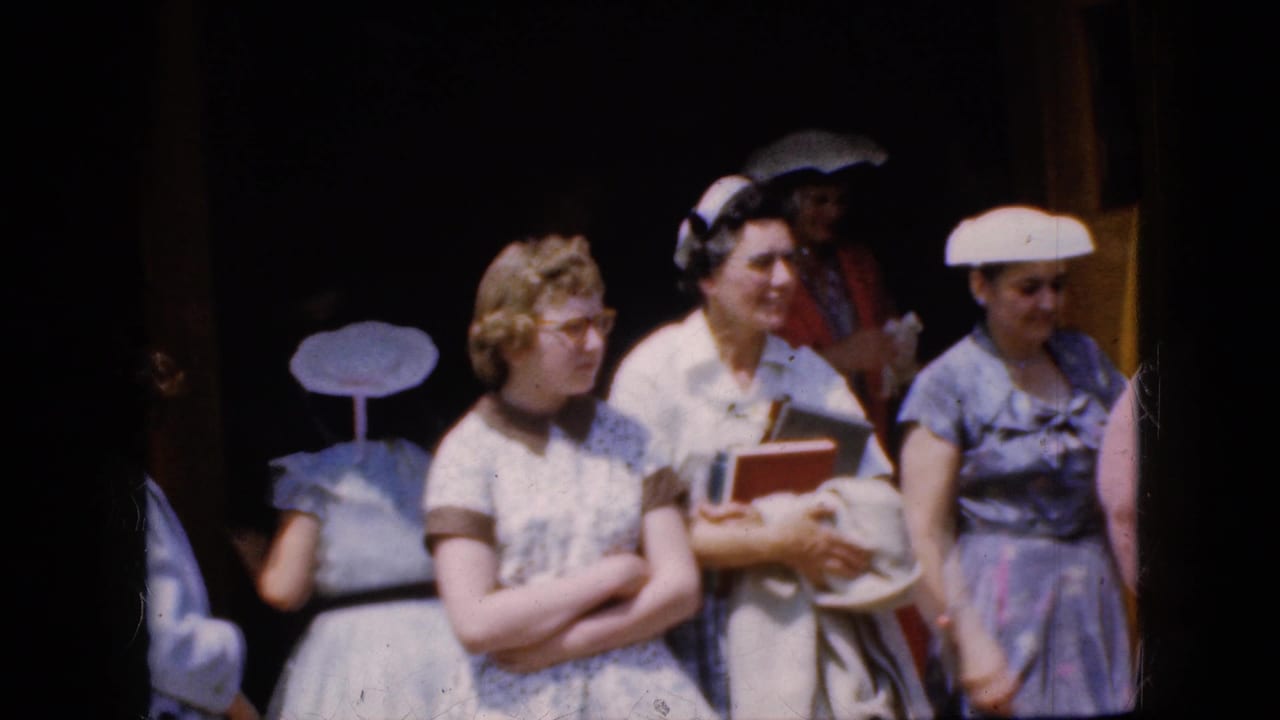 Women in dresses and hats stand in bright sunlight outside a dark doorway.