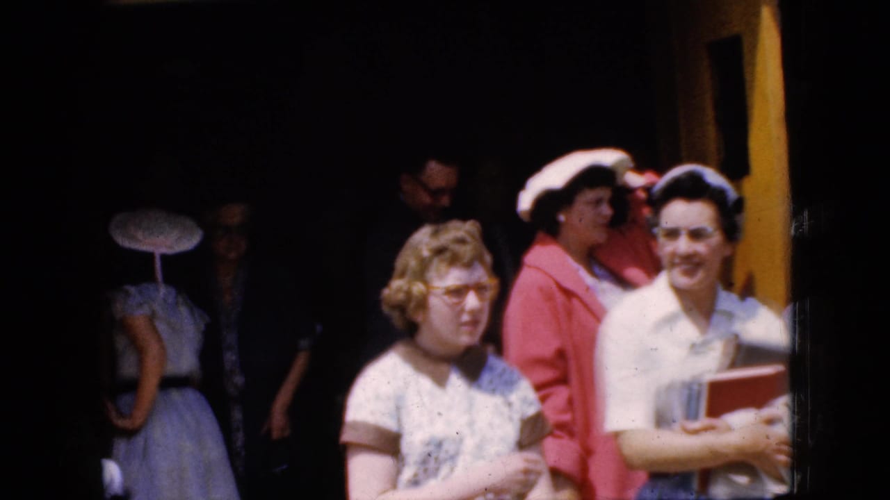 Several women stand near a doorway, including one in a pink coat and another holding red books.