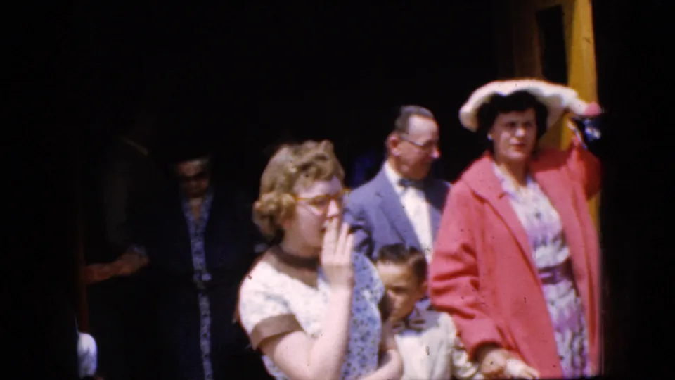 1969 California 8mm frame showing women and a partly visible child outside a church entrance, with a woman in a pink coat and another woman in glasses near the foreground.