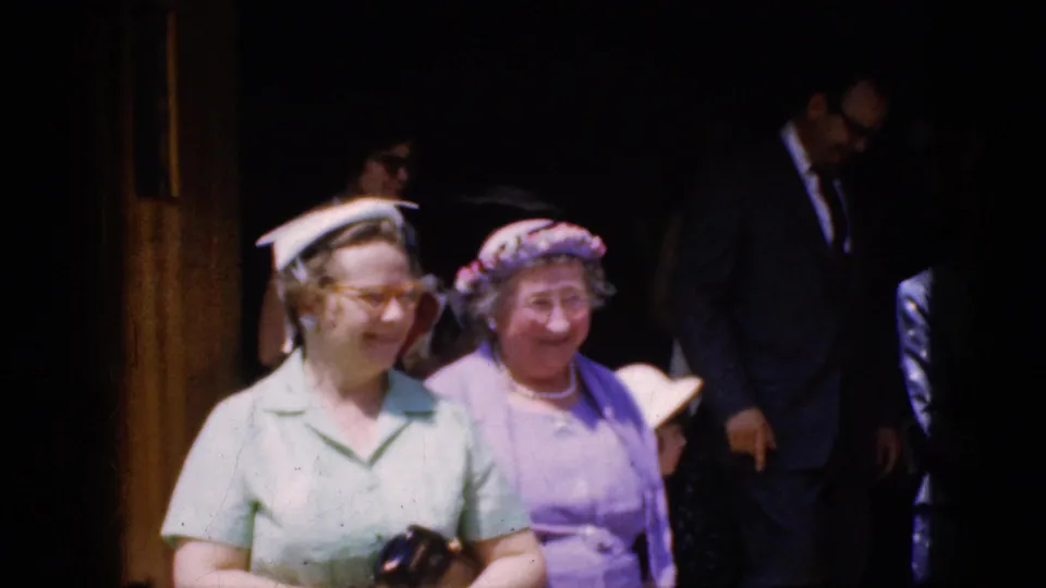 1969 California home-movie frame showing two women in pastel dresses and hats outside a church doorway, with one carrying a small camera in her hands.