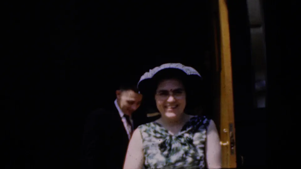 California 1969 8mm frame of a smiling woman in a patterned sleeveless dress and dark hat near a yellow church door, with a man partly visible behind her.