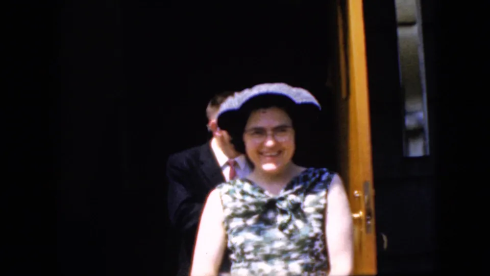California 1969 8mm frame showing a smiling woman in a patterned dress and dark hat at a church entrance, with suited guests partly visible in shadow.