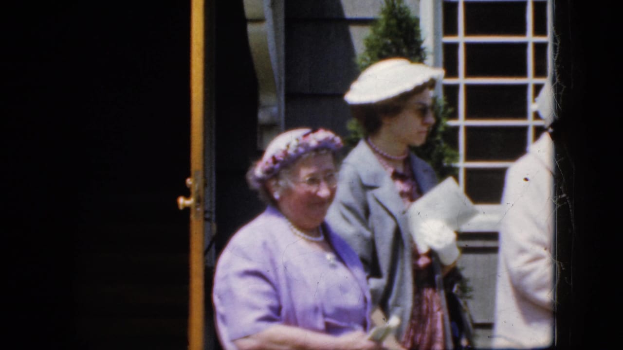 Three adult women in hats and coats stand outside a gray house facade.