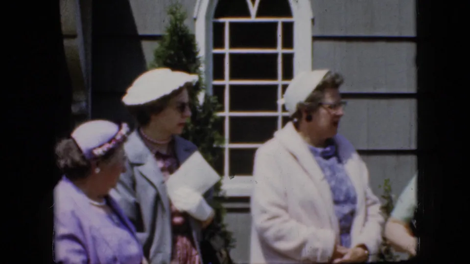 A small group of adult women in hats, coats, and glasses stand in profile outside a house in California home-movie footage from 1969.