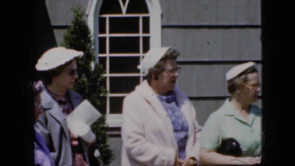 Four adult women in 1969 California home-movie footage stand outside a gray house facade, with hats, coats, eyeglasses, and a small dark camera or case visible.