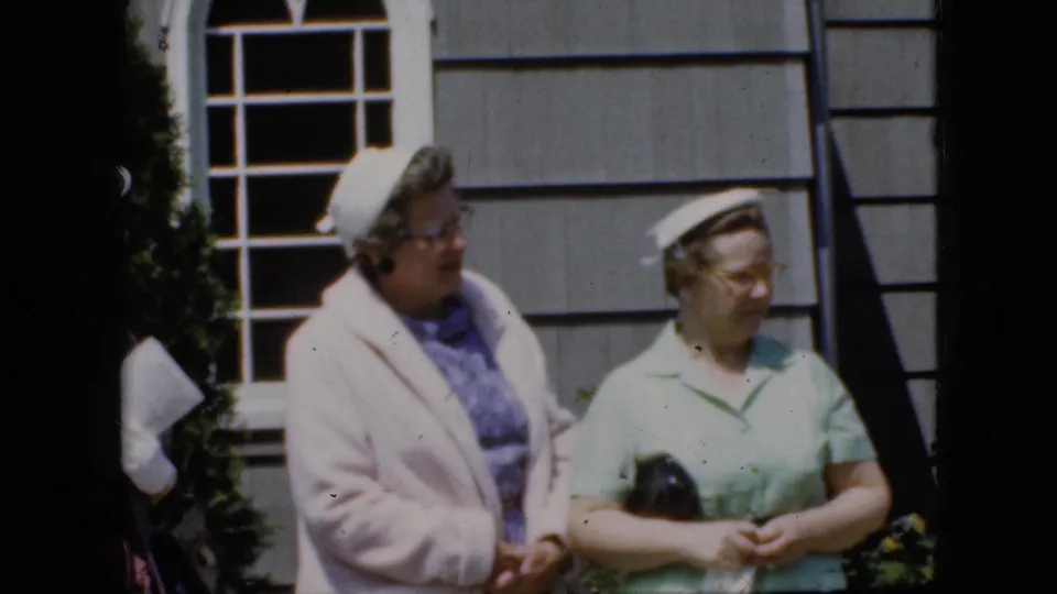 Two older women in late-1960s attire stand outside a gray shingled building, one in a pale pink coat and one in a mint-green top holding a small black purse.