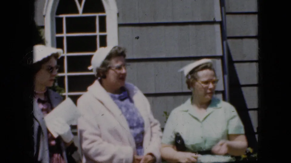 Three older women appear in a 1969 California home-movie frame outside a gray shingled house, with one woman partly entering from the left holding papers.