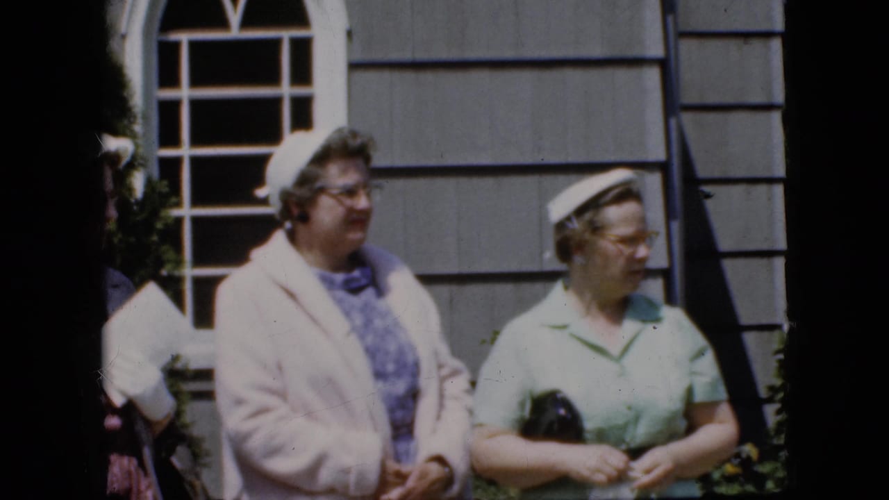 Two women in hats stand outside a gray-sided house near a white-paned window.
