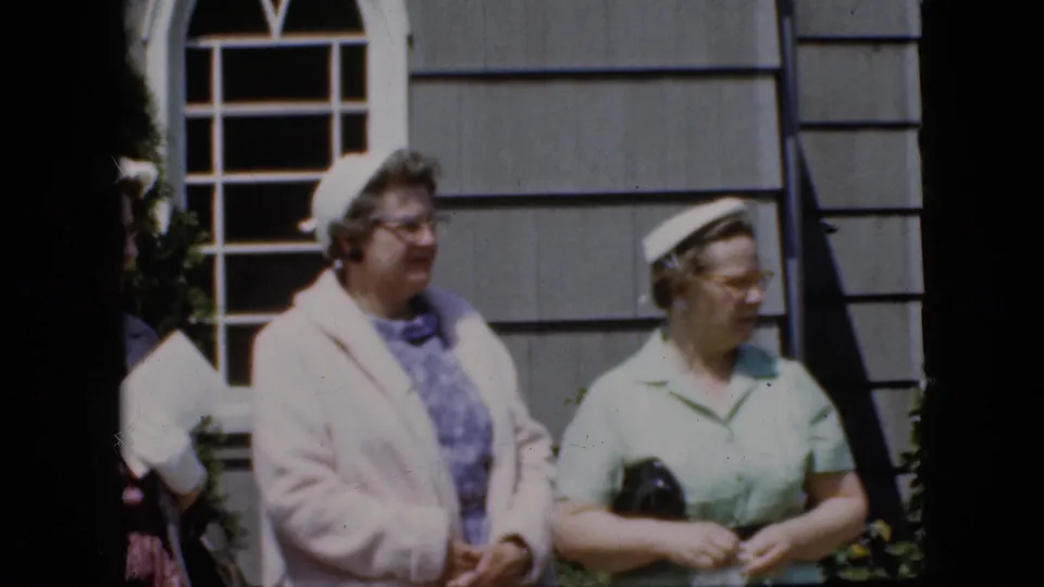 In supported 1969 California home-movie footage, two well-dressed adult women stand outdoors by a gray house wall and white-paned window, one in a pale coat and one in a light green dress holding a dark purse.