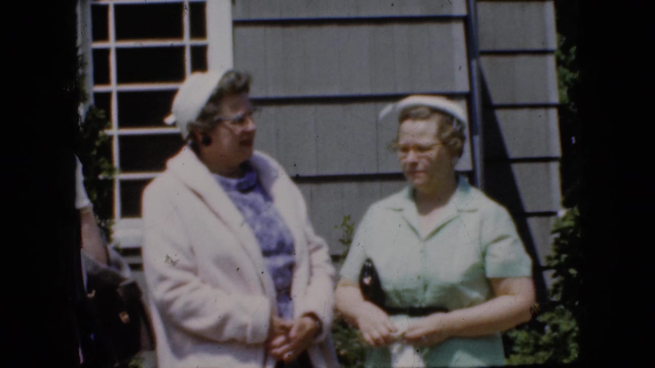 Two women face each other while standing beside a gray house exterior.