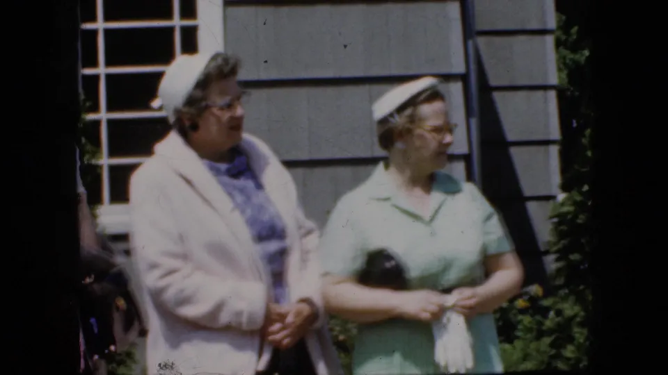 Two adult women in late-1960s attire stand side by side outdoors, with one holding a dark purse and the other wearing a pale coat over a blue patterned dress near a white-paned window.