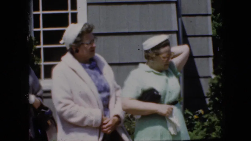 At the end of the 1969 California clip, the woman in green lifts an arm while holding a purse, as the woman in the pale coat stands beside her against the gray house exterior.