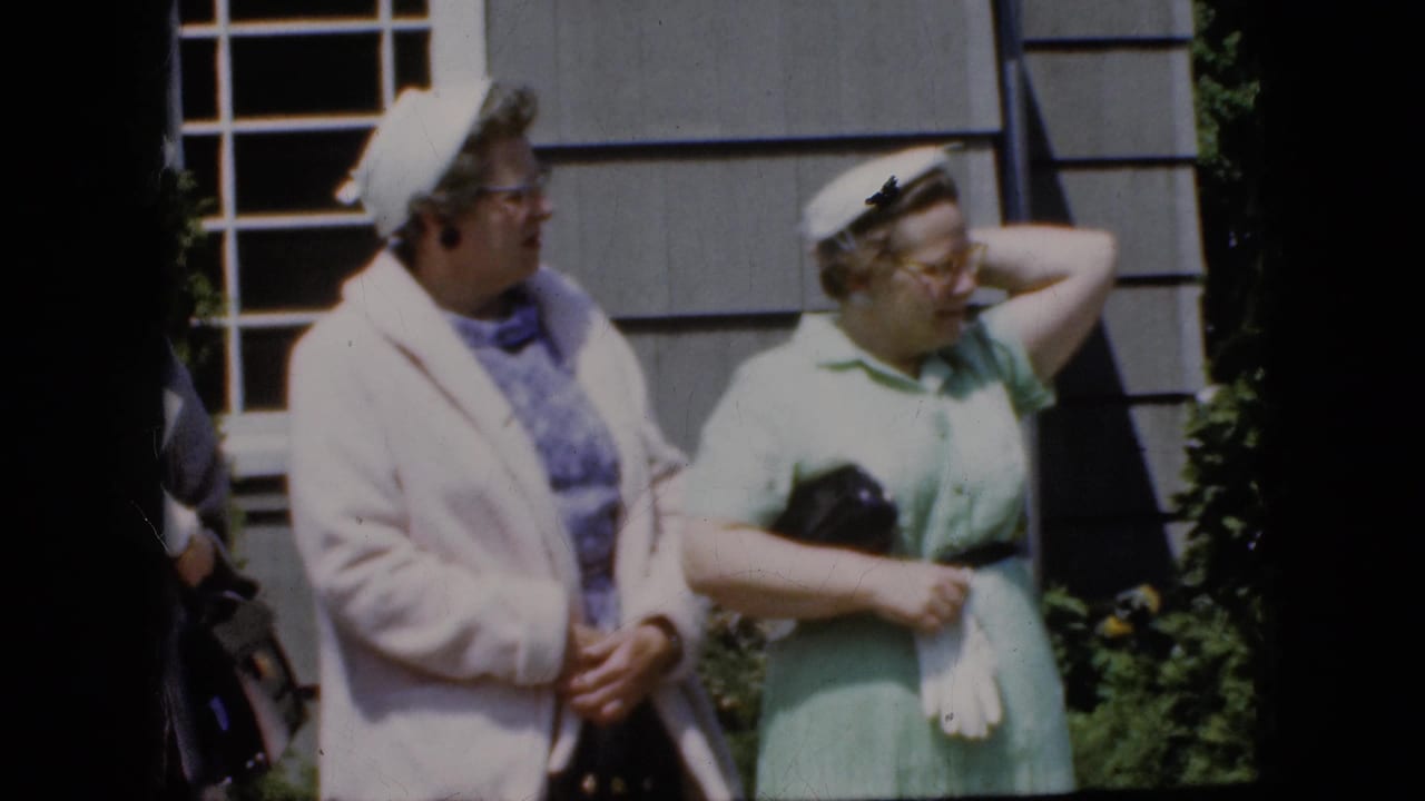 Two adult women in late-1960s attire stand in daylight outside a gray-sided California house, one in a pale coat and one in a light green dress holding gloves.