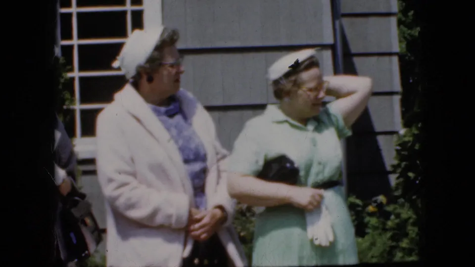 Two adult women in late-1960s attire stand in daylight outside a gray-sided California house, one in a pale coat and one in a light green dress holding gloves.