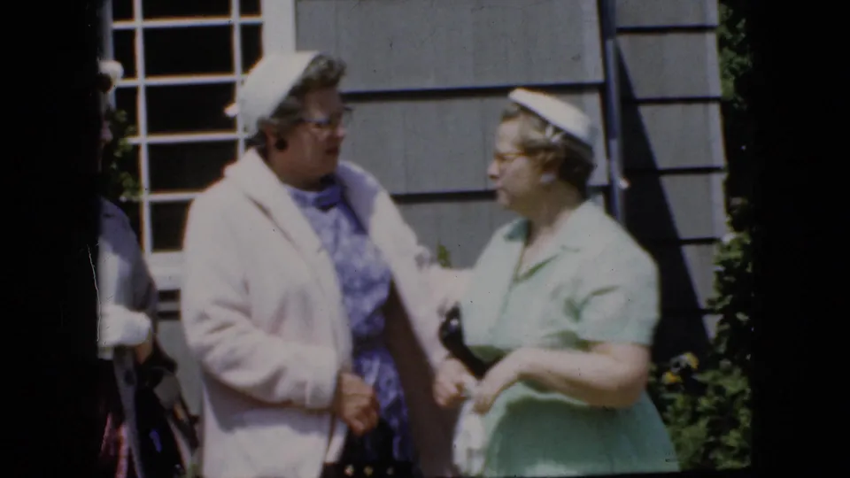 Two adult women face each other outside a gray-sided house in California, with hats, glasses, a pale coat, a green dress, and white gloves visible.