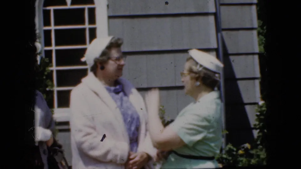 A woman in a light green dress gestures toward a woman in a pale coat outside a gray-sided California house, with film grain and soft focus visible.