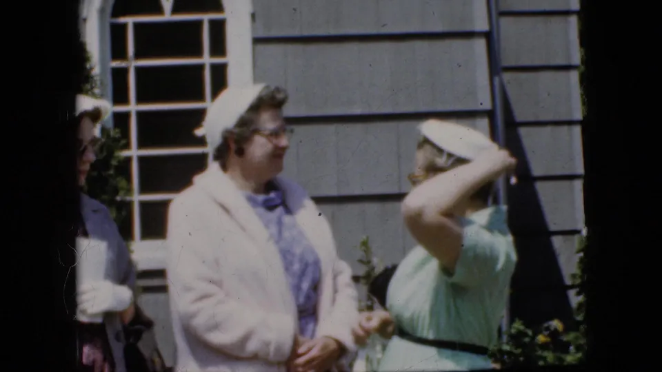 A woman in a light green dress adjusts her white hat beside a woman in a pale coat outside a California house in 1969 home-movie footage.