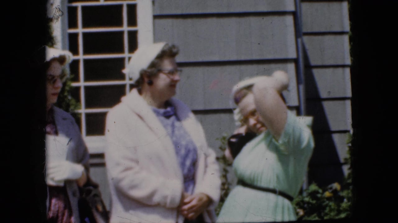 In a 1969 California home-movie frame, adult women in pastel dresses and light jackets stand near a house exterior as one woman raises her arm to adjust her hat.
