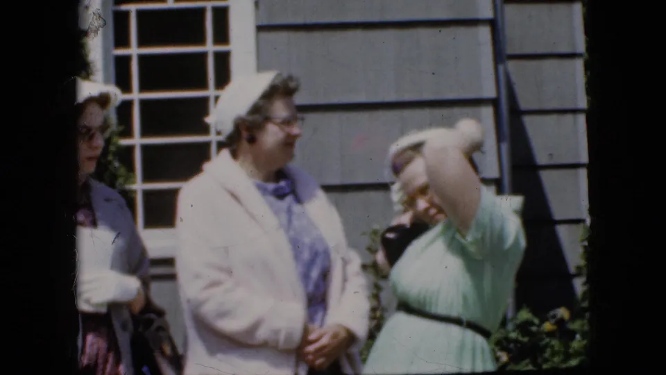In a 1969 California home-movie frame, adult women in pastel dresses and light jackets stand near a house exterior as one woman raises her arm to adjust her hat.