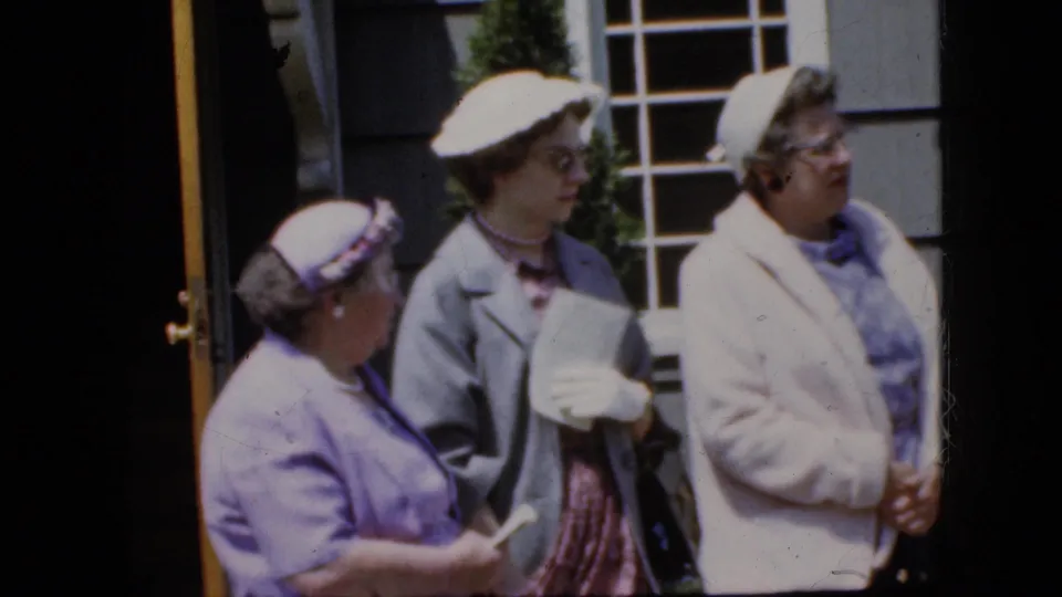 Adult women in coordinated pastel dresses, hats, and coats stand close together outside a house, with one holding papers in the 1969 California home-movie scene.