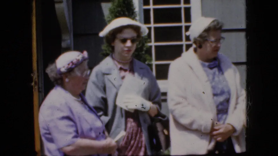The frame shows three adult women in late-1960s pastel formalwear outside a California house, including hats, gloves, papers, eyeglasses, and a dark handbag.