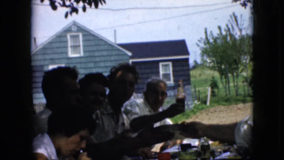 1969 California backyard gathering frame with several adults in partial silhouette at a food-filled table, one person holding a bottle up against a bright yard and house backdrop.