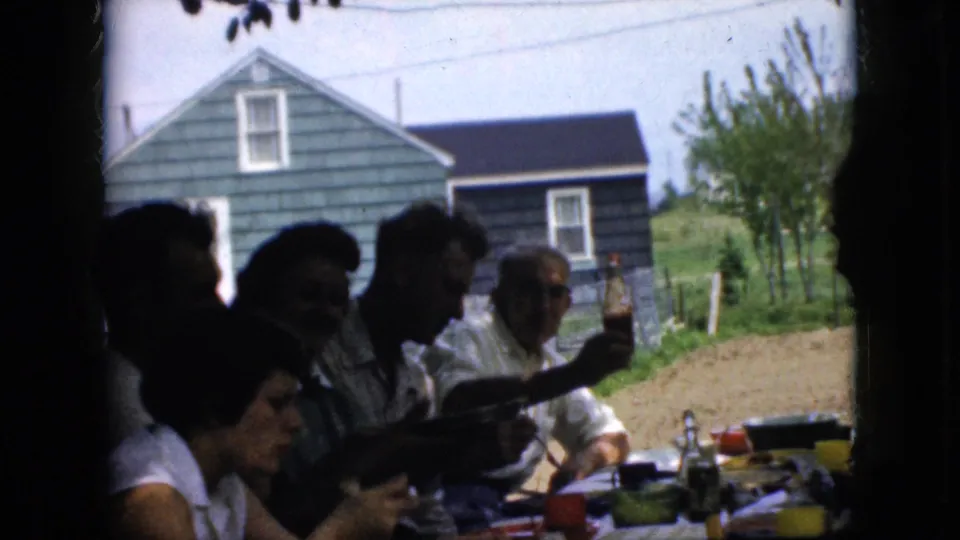 1969 California home-movie frame showing family members eating at a crowded outdoor table, with plates, bottles, bowls, a dirt yard, trees, and a blue-sided house visible beyond them.