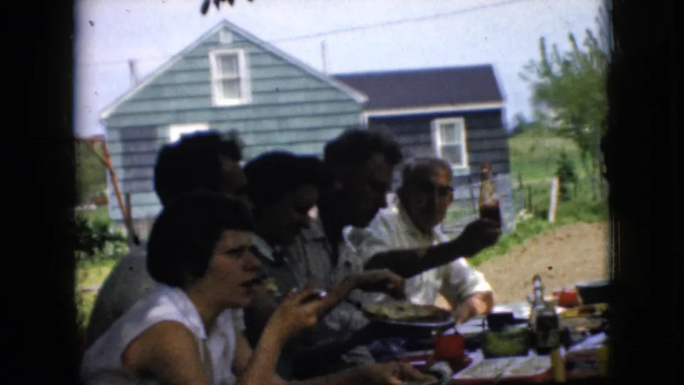 1969 California outdoor meal frame showing a woman eating in the foreground, other adults seated beside her, tableware and food on the table, and a blue house in the background.