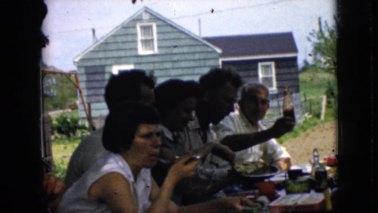 Adults sit at an outdoor table eating, with a green-sided house visible beyond the window frame.