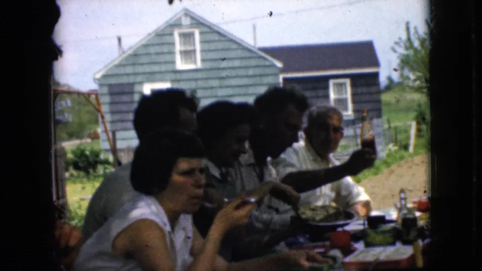 1969 California home-movie frame showing adults gathered at an outdoor table with food and drinks, filmed through a dark window or doorway frame with a green-sided house in the background.