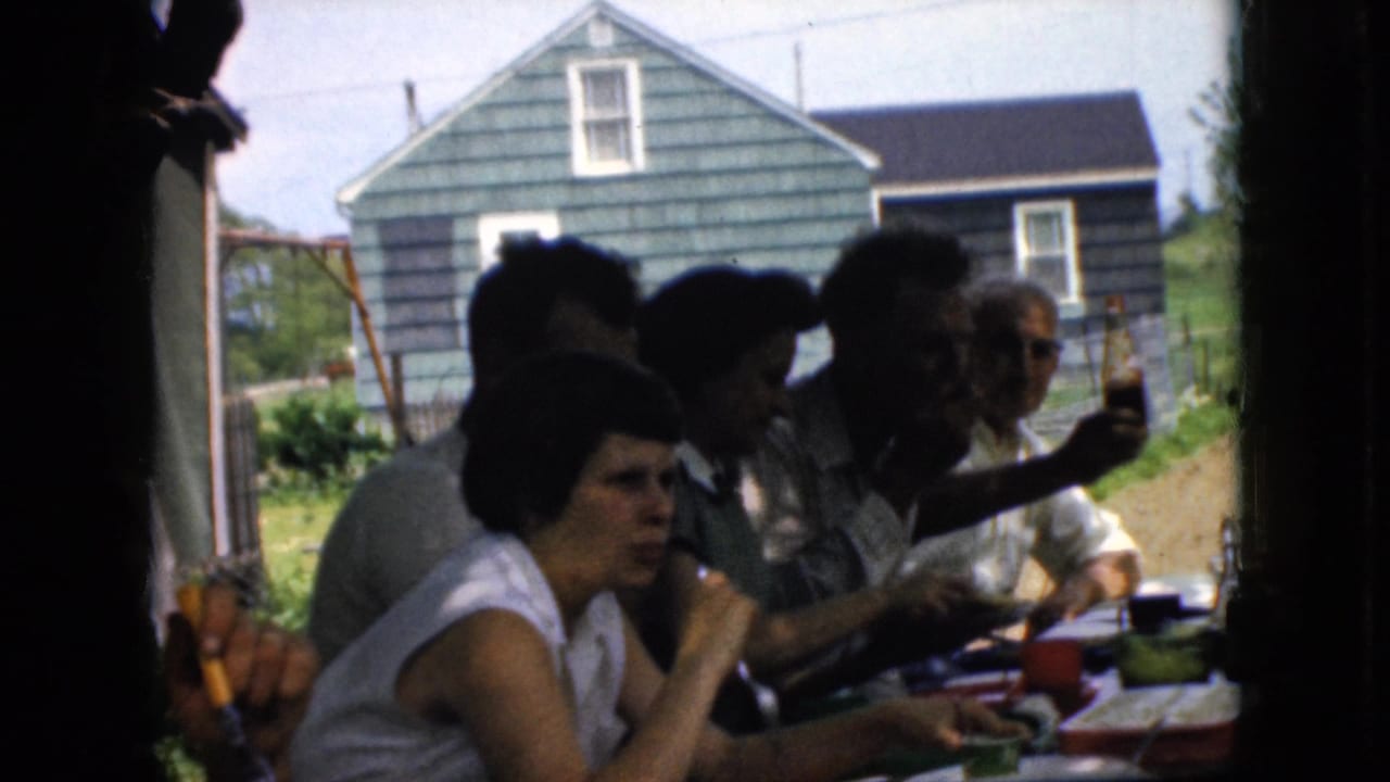 Adults eat and drink at a table while one man raises a bottle near a green-sided house.